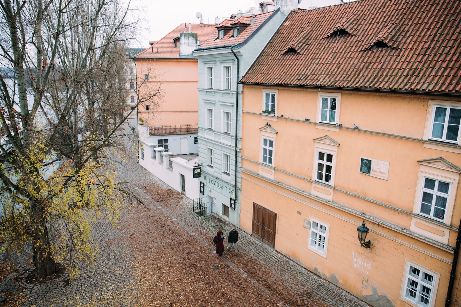גבר ואישה צועדים מחוץ לבית בן 3 קומות ליד עץ ירוק בפראג